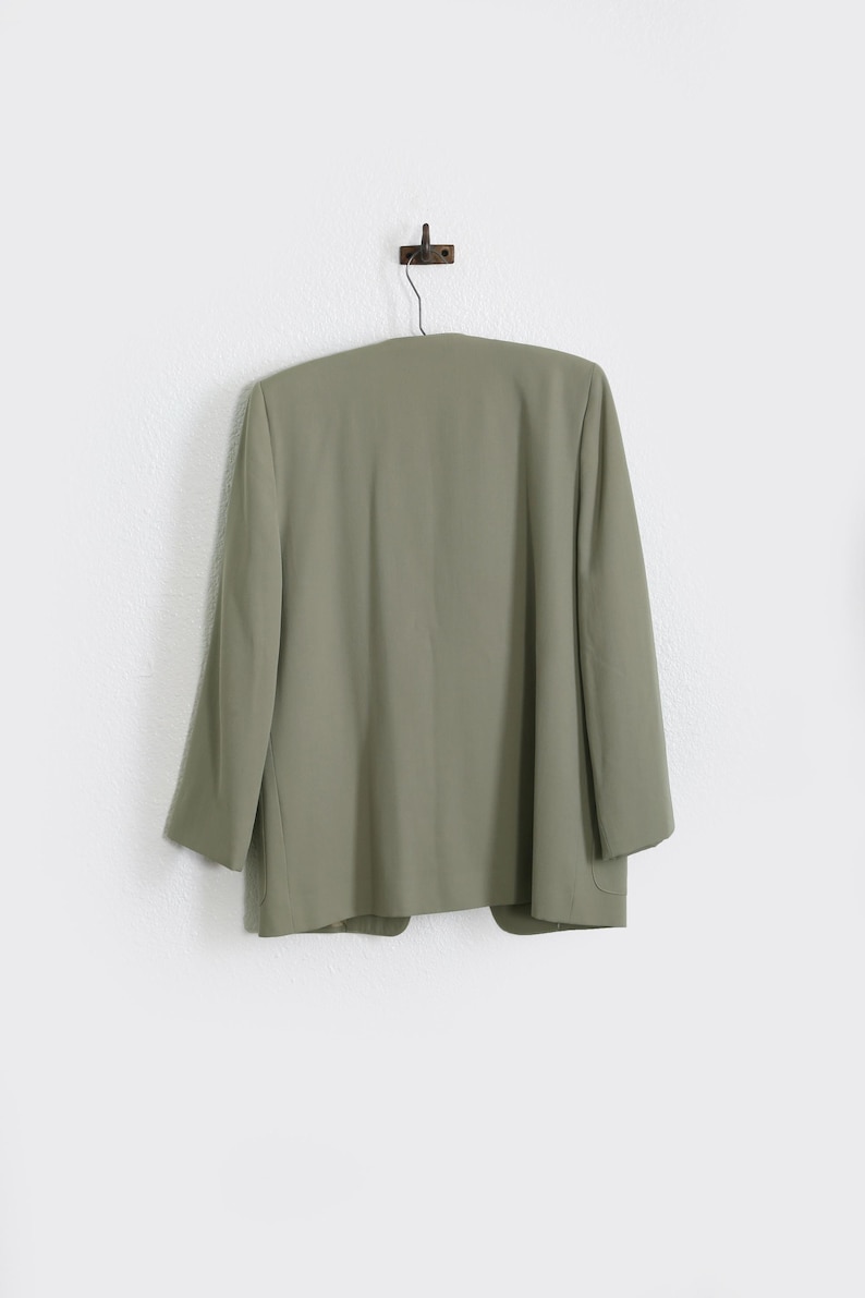 K&ouml;nnte beinhalten: Salbeigr&uuml;ner Blazer, der an einer Wand h&auml;ngt. Der Blazer hat ein klassisches Design mit langen &Auml;rmeln und einer taillierten Passform. Die Farbe ist ein ged&auml;mpfter, erdiger Ton, und das Kleidungsst&uuml;ck scheint aus einem glatten Stoff zu bestehen. Der Blazer h&auml;ngt an einem dunklen Metallhaken.