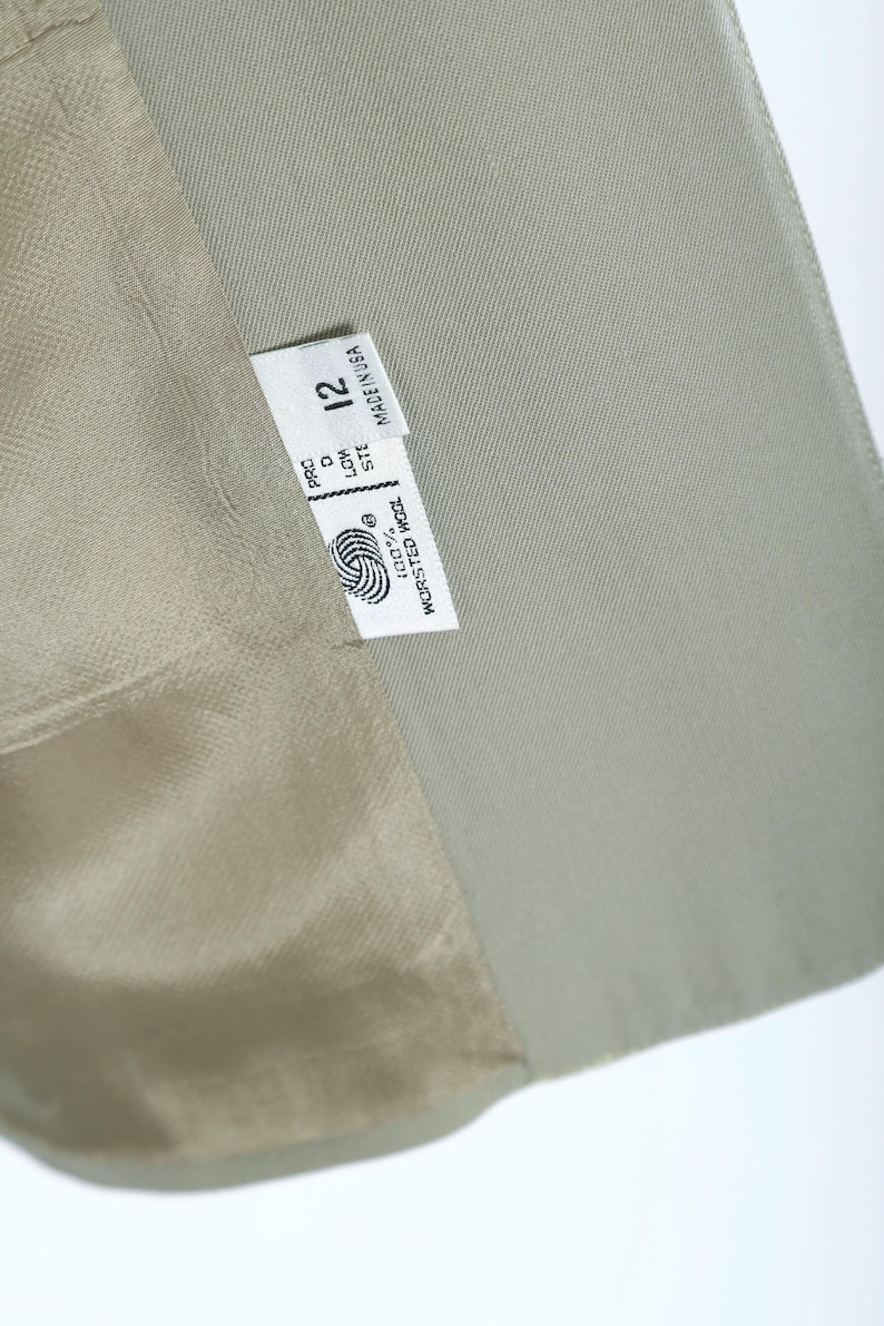 K&ouml;nnte beinhalten: Nahaufnahme eines hellgr&uuml;nen Blazers mit einem Etikett mit der Aufschrift "Made in USA" und der Gr&ouml;&szlig;e "12". Das Etikett enth&auml;lt auch ein Wollsymbol und die Worte "100% Worsted Wool". Das Innenfutter des Blazers ist hellbeige.