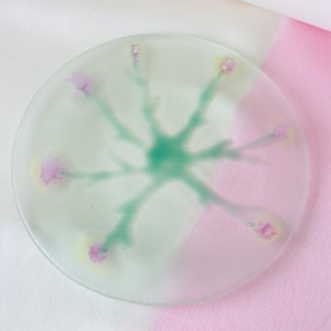 May include: A round, frosted glass plate with a floral design. The design features a central green burst with radiating stems ending in pink and yellow flower-like shapes. The plate rests on a white and pink surface.