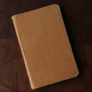 Pode incluir: Um livro de campo vintage castanho claro com texto em relevo. A capa diz "ALCO FIELD BOOK 760". O livro tem formato retangular e um aspeto ligeiramente desgastado. O fundo é uma superfície de madeira escura. O design sugere um item antigo.