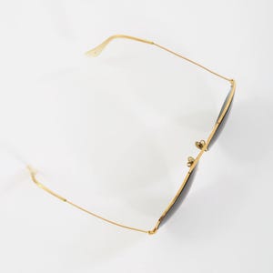 May include: A pair of sunglasses with thin gold-tone metal frames and dark, tinted lenses. The eyewear features a classic aviator-style silhouette with slender temples, a double bridge, and adjustable nose pads, captured from a high angle.