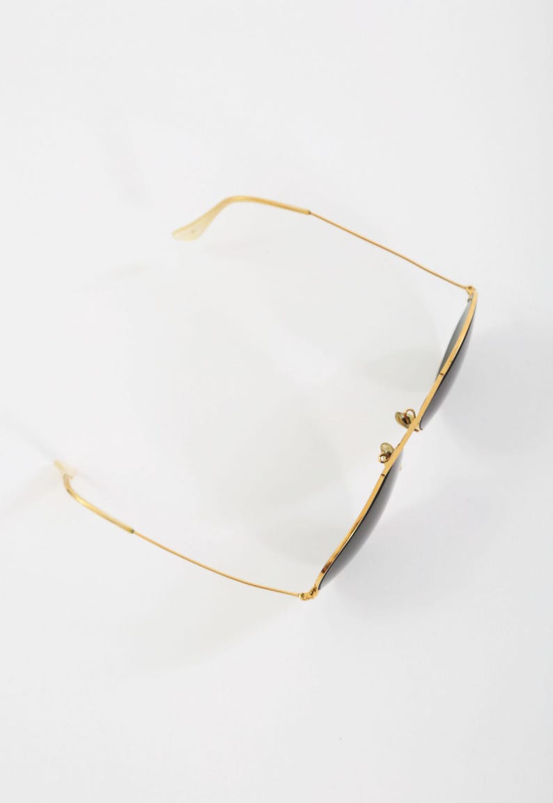 May include: A pair of sunglasses with thin gold-tone metal frames and dark, tinted lenses. The eyewear features a classic aviator-style silhouette with slender temples, a double bridge, and adjustable nose pads, captured from a high angle.