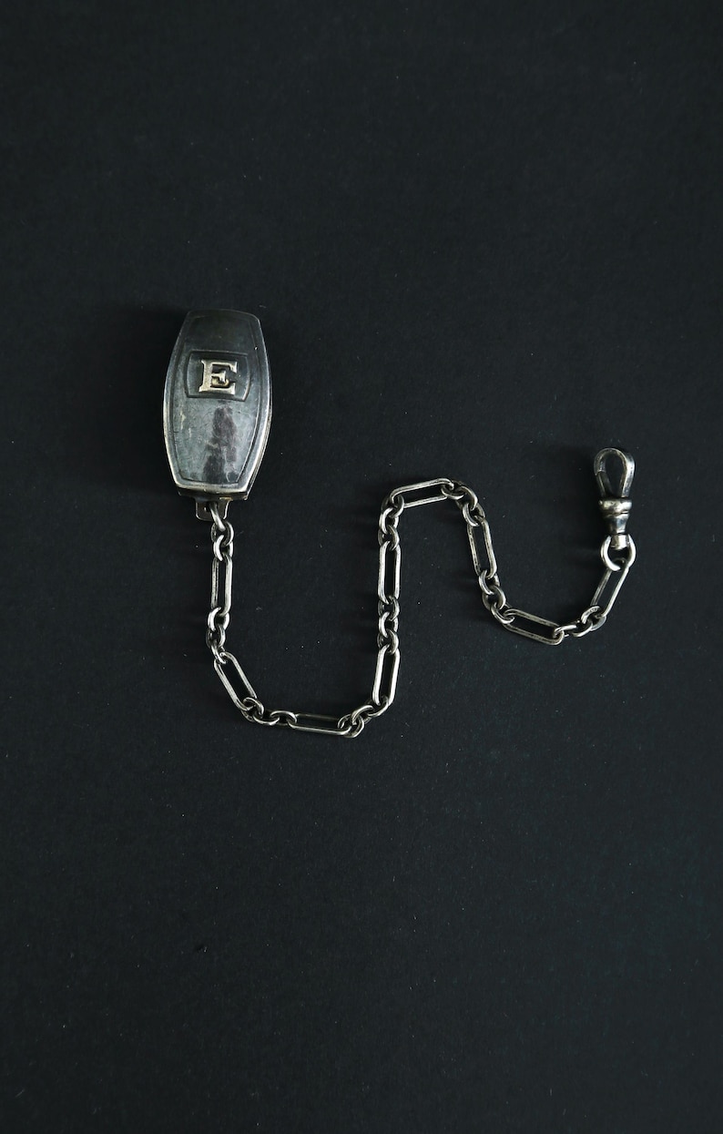 May include: A silver pocket watch chain with a rectangular fob featuring the letter "E". The chain is composed of elongated links and terminates in a clasp. The fob and chain are set against a black background.