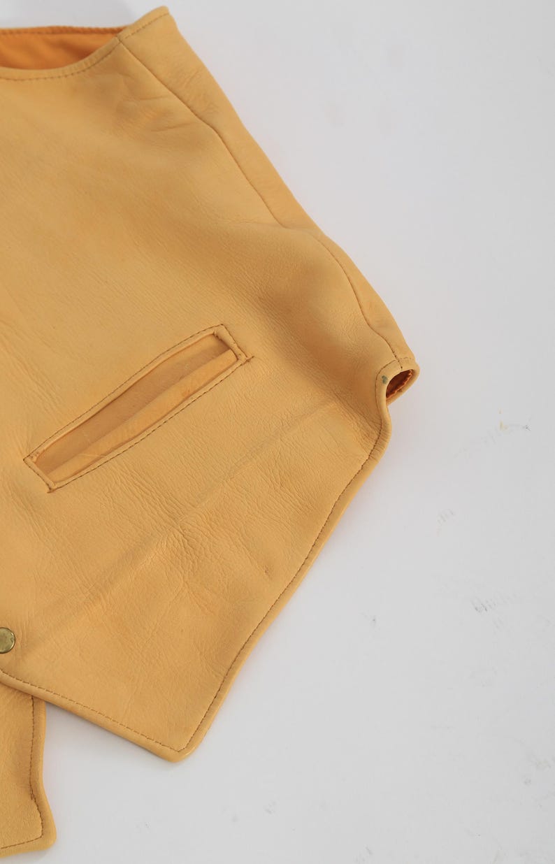 May include: A yellow leather vest with a pocket on the front. The vest has a pointed collar and a button closure.
