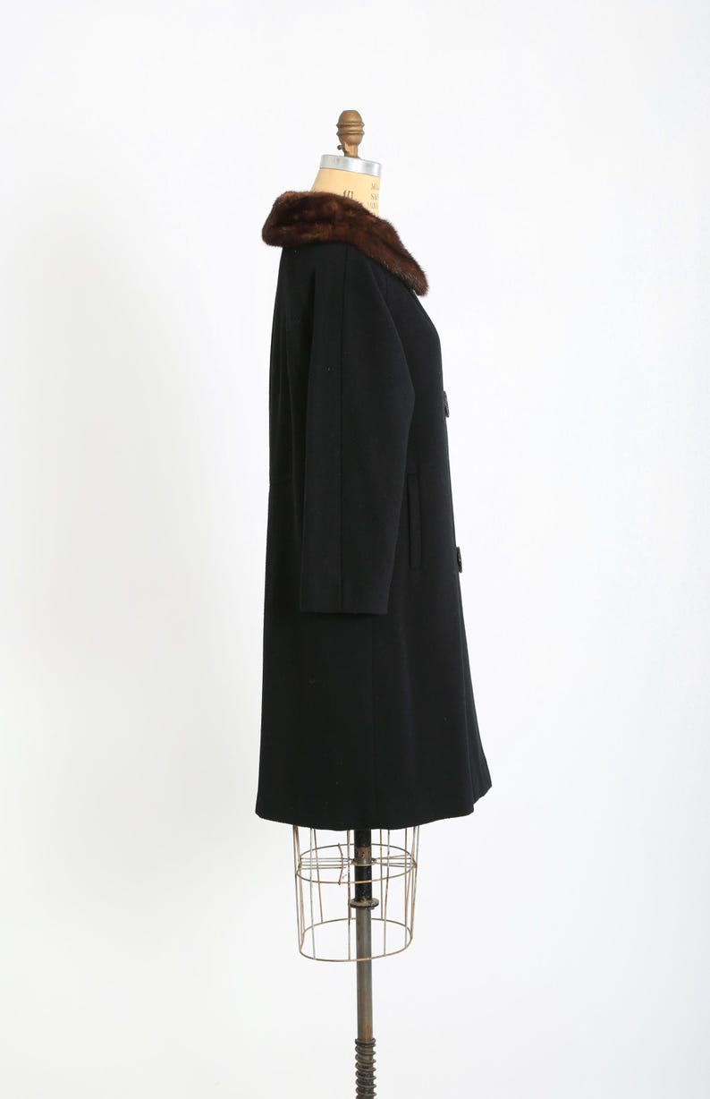 May include: A vintage black coat with a brown fur collar. The coat is displayed on a mannequin and features a classic design with a button closure. The coat's length extends to the mid-calf, offering a timeless silhouette.