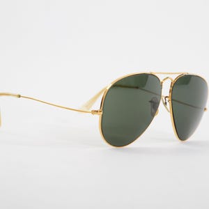 May include: Aviator sunglasses featuring a thin gold metal frame and dark green teardrop-shaped lenses. The design includes a double bridge across the top and slender temples with clear plastic tips, providing a classic look with a metallic finish.