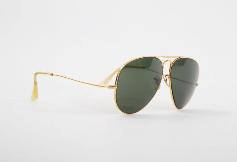 May include: Aviator sunglasses featuring a thin gold metal frame and dark green teardrop-shaped lenses. The design includes a double bridge across the top and slender temples with clear plastic tips, providing a classic look with a metallic finish.