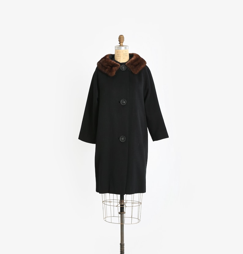 May include: A vintage black coat with a brown fur collar and three large black buttons. The coat is displayed on a dress form against a white background. The coat has long sleeves and a classic silhouette.