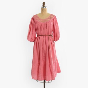 K&ouml;nnte beinhalten: Ein knielanges rosa Kleid mit dreiviertellangen Puff&auml;rmeln und einem Rundhalsausschnitt mit vertikalen Ausschnittdetails. Das Kleid hat eine geraffte Taille, die mit einem d&uuml;nnen braunen Kordelg&uuml;rtel gebunden ist, und einen gestuften Rock.
