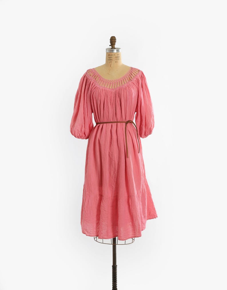 K&ouml;nnte beinhalten: Ein knielanges rosa Kleid mit dreiviertellangen Puff&auml;rmeln und einem Rundhalsausschnitt mit vertikalen Ausschnittdetails. Das Kleid hat eine geraffte Taille, die mit einem d&uuml;nnen braunen Kordelg&uuml;rtel gebunden ist, und einen gestuften Rock.