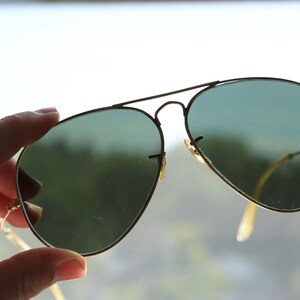 May include: Aviator sunglasses featuring a thin gold-tone metal frame and teardrop-shaped green tinted lenses. The eyewear includes a double bridge design across the top and slender metal temples with curved ends for a secure fit over the ears.
