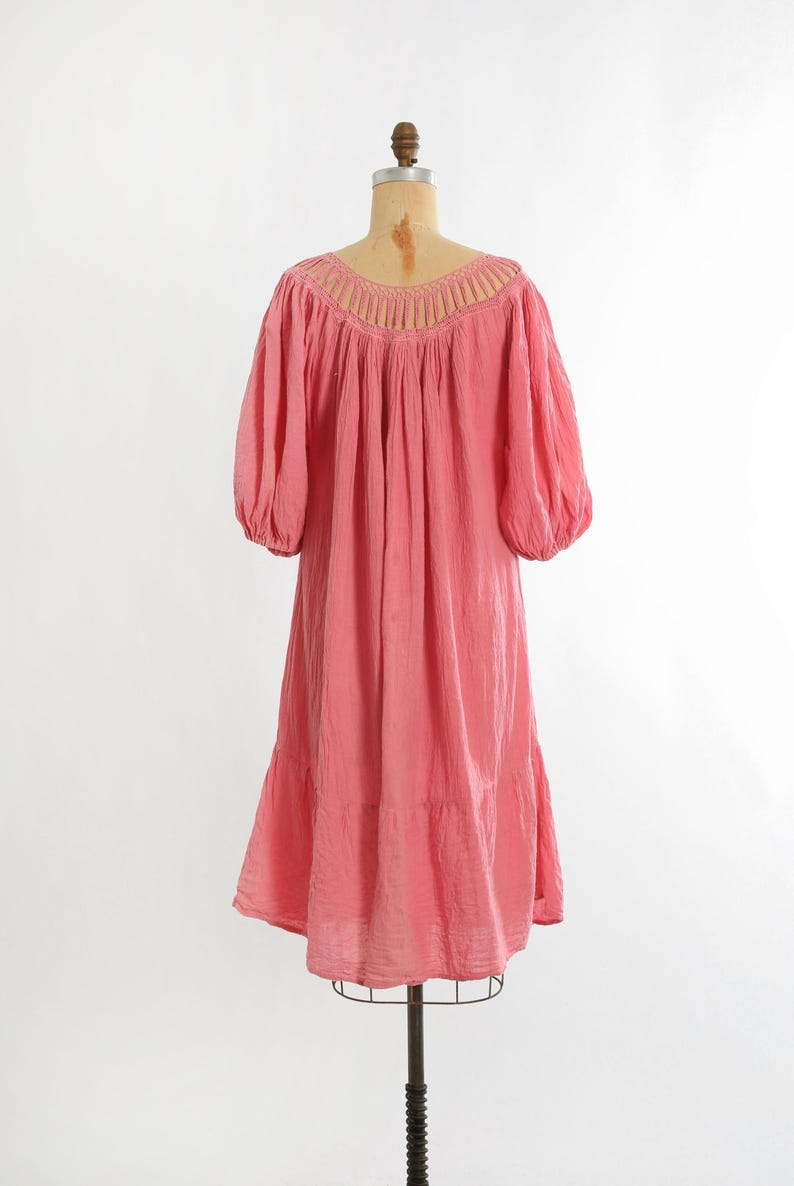 K&ouml;nnte beinhalten: Ein korallrosa Kleid im Vintage-Stil mit einem Rundhalsausschnitt und kurzen, gepufften &Auml;rmeln. Das Kleid hat einen Stufenrock und wird auf einer Schaufensterpuppe pr&auml;sentiert. Der Stoff wirkt leicht und flie&szlig;end.