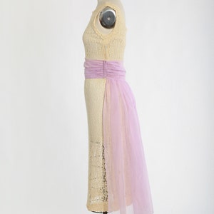 Op de afbeelding: Een vintage cr&egrave;mekleurige kanten jurk met een lavendelkleurige tule overlay en een bijpassende band. De jurk heeft een getailleerd lijfje en een zwierige rok.