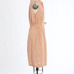 May include: A pink and orange checked sleeveless dress with a button-up front and a tie at the waist. The dress has a single pocket on the right side.