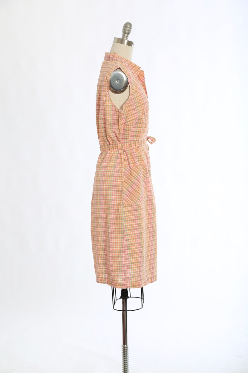 May include: A pink and orange checked sleeveless dress with a button-up front and a tie at the waist. The dress has a single pocket on the right side.