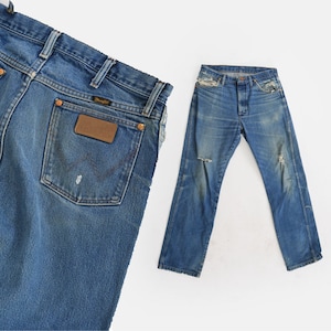 Op de afbeelding: Twee paar blauwe denim jeans. De jeans aan de linkerkant hebben een bruin leren patch met het woord "Wrangler" erop. De jeans aan de rechterkant hebben een lichtblauwe wassing en zijn licht beschadigd.