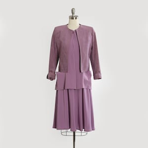 Puede incluir: Conjunto de vestido de dos piezas lavanda vintage sobre un maniquí. El conjunto incluye una chaqueta de manga larga con bolsillos y cierre de botón, y un vestido a juego hasta la rodilla. El vestido tiene una falda plisada y un corpiño en capas.