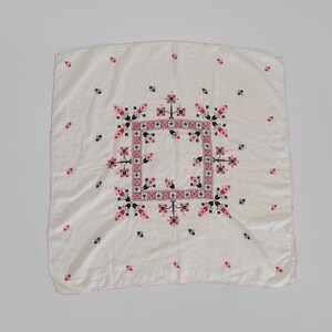May include: A white linen tablecloth with a red and black embroidered design. The design features a central square with a geometric pattern surrounded by stylized floral motifs.
