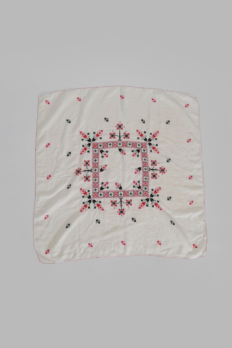 May include: A white linen tablecloth with a red and black embroidered design. The design features a central square with a geometric pattern surrounded by stylized floral motifs.