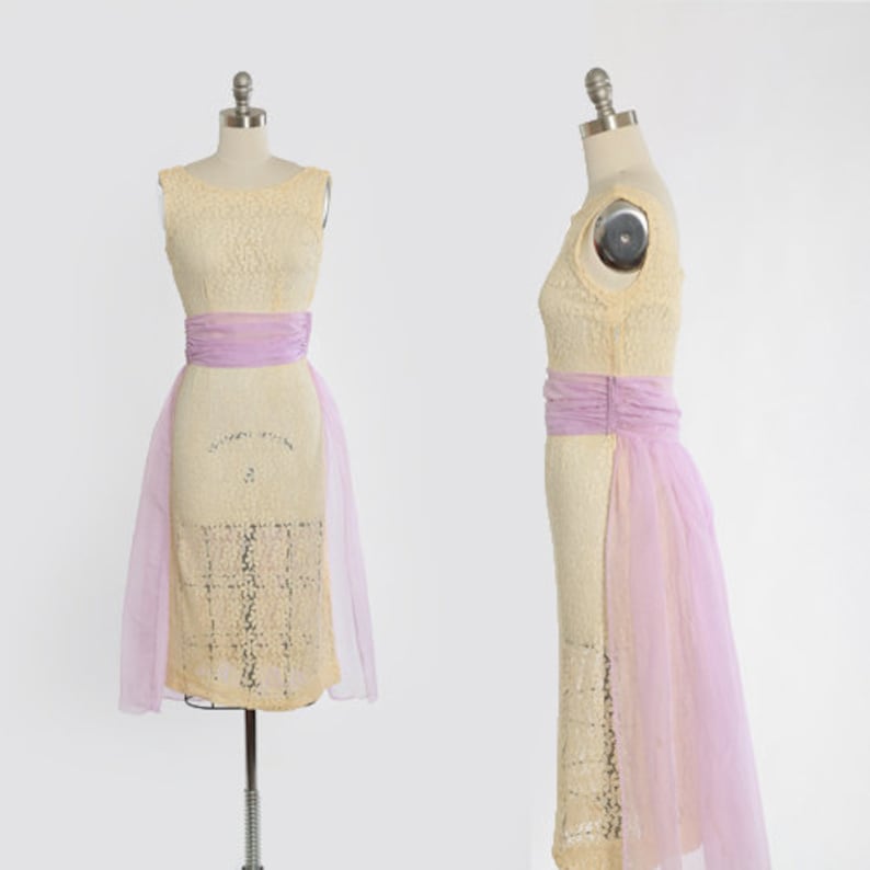Op de afbeelding: Een mouwloze, cr&egrave;mekleurige kantenjurk met een bijpassende transparante overlay en een lavendel satijnen ceintuur. De jurk heeft een aansluitend lijfje en een volle rok.