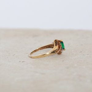 May include: A gold ring featuring a rectangular emerald-cut green gemstone. The ring has a twisted design with small accent stones on the side. The ring is set on a neutral surface.