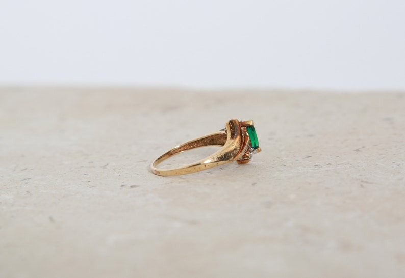 May include: A gold ring featuring a rectangular emerald-cut green gemstone. The ring has a twisted design with small accent stones on the side. The ring is set on a neutral surface.