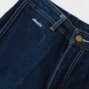 May include: Close-up of dark blue denim pants with white stitching. The pants have a brand label with the word "chardon" embroidered on the back waistband.