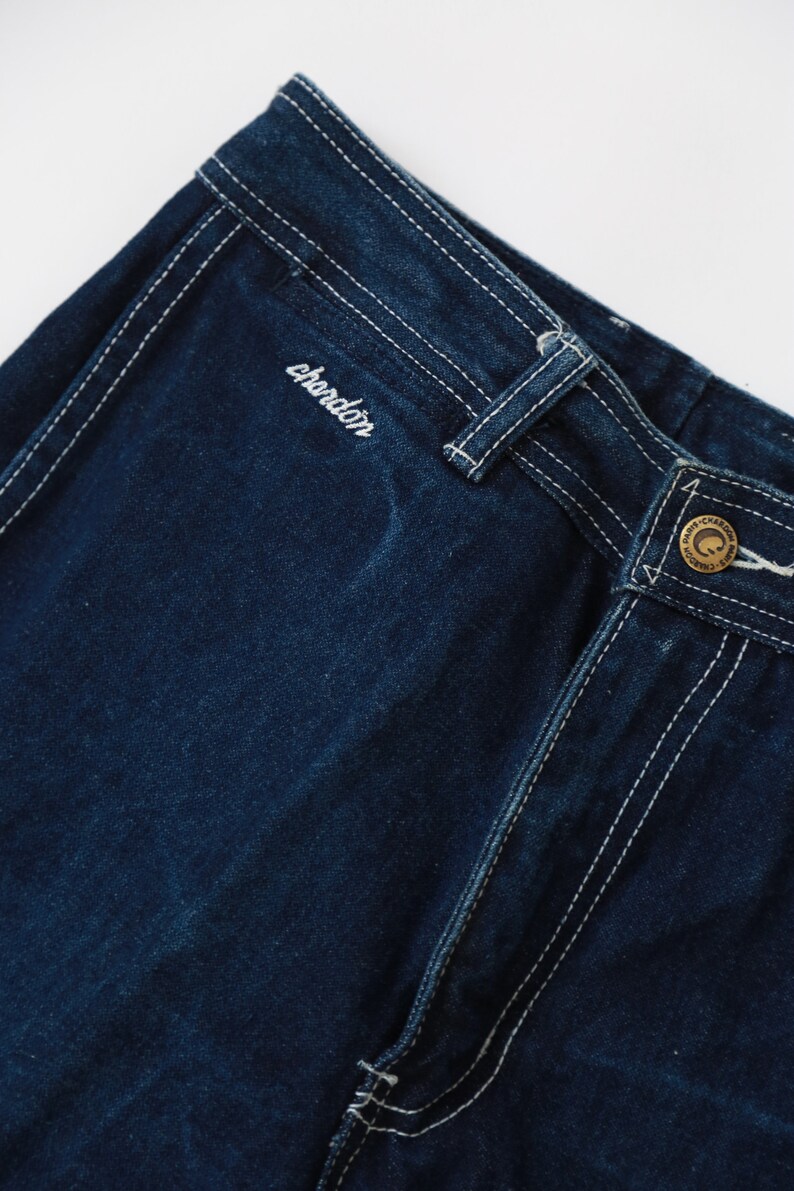 May include: Close-up of dark blue denim pants with white stitching. The pants have a brand label with the word "chardon" embroidered on the back waistband.