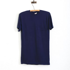 May include: A navy blue short-sleeved t-shirt with a chest pocket hangs from a wooden hanger. The shirt is made of a soft material and is displayed against a white background. The t-shirt is a solid colour.