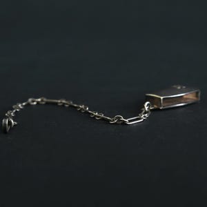 May include: A silver-toned matchbox holder with a chain, likely for a pocket or purse. The rectangular holder has a hinged lid and is connected to a delicate chain. The design is simple and elegant, suitable for everyday use.