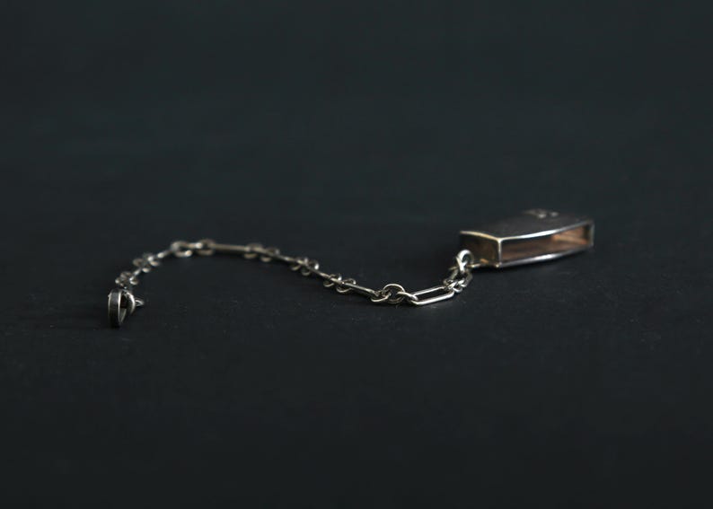 May include: A silver-toned matchbox holder with a chain, likely for a pocket or purse. The rectangular holder has a hinged lid and is connected to a delicate chain. The design is simple and elegant, suitable for everyday use.