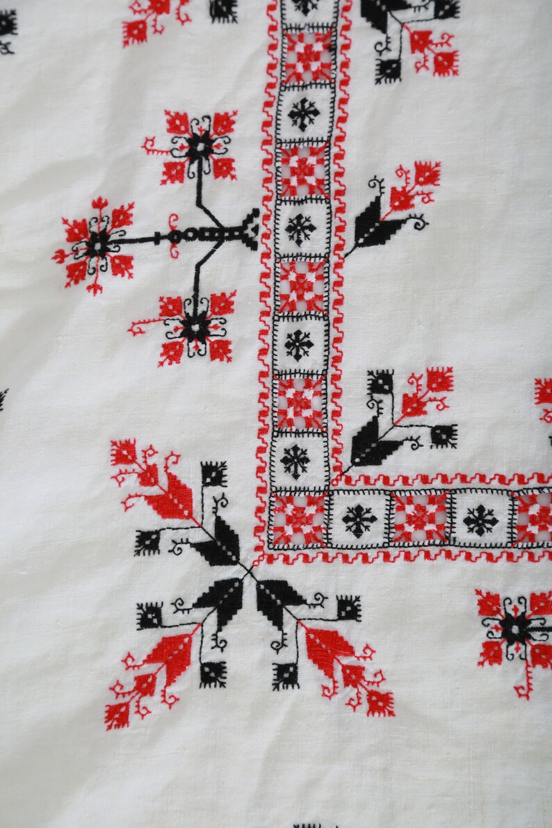 May include: White fabric with a black and red embroidered design. The design features a repeating pattern of geometric shapes and stylized flowers.