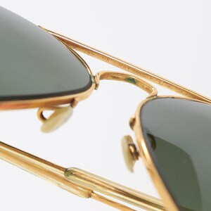 May include: Gold-tone aviator sunglasses featuring dark green lenses and a double bridge design. The metal frame includes adjustable nose pads and thin temples. The top bar of the bridge is engraved with text that is partially visible.
