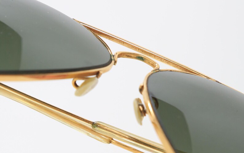 May include: Gold-tone aviator sunglasses featuring dark green lenses and a double bridge design. The metal frame includes adjustable nose pads and thin temples. The top bar of the bridge is engraved with text that is partially visible.