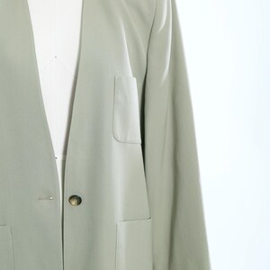 K&ouml;nnte beinhalten: Ein hellgr&uuml;ner Blazer mit Reverskragen, zwei Vordertaschen und einem einzelnen Knopfverschluss. Der Blazer wird auf einer Schaufensterpuppe pr&auml;sentiert, die seine ma&szlig;geschneiderte Passform und sein Design hervorhebt. Die &Auml;rmel sind lang und der Stoff scheint leicht zu sein.