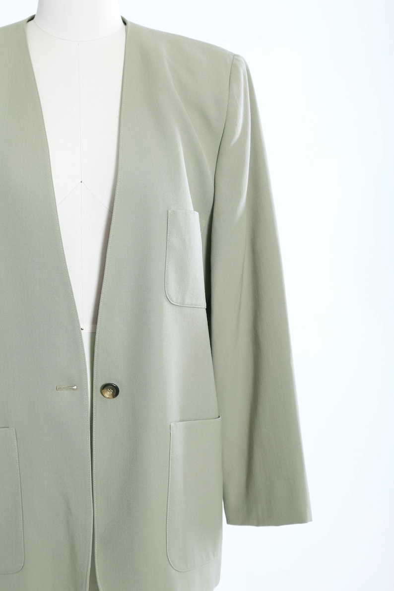 K&ouml;nnte beinhalten: Ein hellgr&uuml;ner Blazer mit Reverskragen, zwei Vordertaschen und einem einzelnen Knopfverschluss. Der Blazer wird auf einer Schaufensterpuppe pr&auml;sentiert, die seine ma&szlig;geschneiderte Passform und sein Design hervorhebt. Die &Auml;rmel sind lang und der Stoff scheint leicht zu sein.