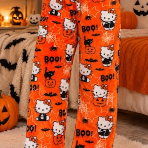 Puede incluir: Pantalones de pijama de forro polar naranja brillante con un estampado de Halloween de personajes de Hello Kitty, gatos negros, calabazas, telarañas, estrellas y murciélagos, con el texto "BOO!" impreso en negro por toda la tela.