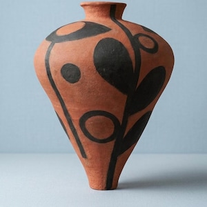 Puede incluir: Un jarrón de color terracota con una base cónica y estrecha y un cuello fino. La superficie presenta motivos botánicos abstractos pintados a mano en negro, incluyendo formas circulares y figuras similares a hojas a lo largo del cuerpo.