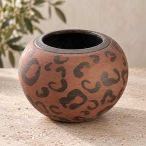 May include: A round, terracotta-coloured ceramic bowl featuring a dark charcoal leopard print pattern across its exterior surface. The vessel has a wide, open top with a thick, dark-toned rim and a deep, dark interior, showcasing a matte finish.