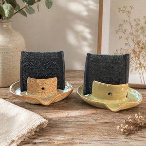 Puede incluir: Dos soportes de cerámica para esponjas con acabado brillante, uno en color beige moteado y otro en verde claro. Cada soporte presenta una pieza curva con un pequeño orificio circular, diseñada para sostener una esponja negra rectangular.