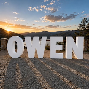 May include: Large, freestanding white block letters spelling the name OWEN, positioned on a gravel surface. The letters are solid, matte, and uppercase, standing against a backdrop of mountains, a lake, and a sunset sky with scattered clouds.