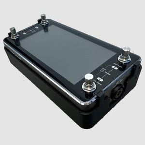 May include: A black rectangular electronic effects pedal featuring a large central touchscreen display, four metallic footswitches at the corners, and printed labels including Hold, Modes, and Hold: Tempo. The device has a sleek, dark finish.