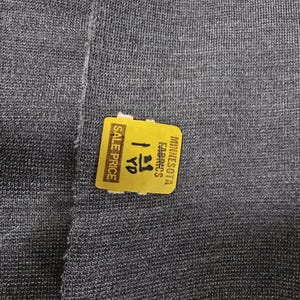 Könnte beinhalten: Ein Stück dunkelgrauer Stoff mit einer feinen, gleichmäßigen Strickstruktur. Auf der Oberfläche befindet sich ein gelber rechteckiger Aufkleber mit dem Text "MINNESOTA FABRICS" und "SALE PRICE" sowie der handschriftlichen Angabe "1 3/8 YD".