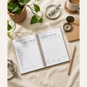 May include: An open spiral-bound notebook featuring a Plant Mapping Grid with columns for plant names and care tracking. The white pages contain black printed text and illustrations of various plants, with a wooden pencil resting nearby.
