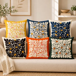 May include: A collection of six square throw pillows arranged on a sofa. Each pillow features a repeating daisy flower pattern in various color combinations, including yellow, navy blue, black, orange, and dark green with white petals.