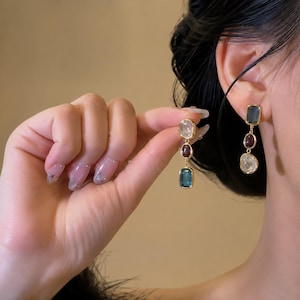 Puede incluir: Pendientes colgantes con tres piedras alineadas verticalmente en engastes de tono dorado. La piedra superior es de color gris azulado translúcido, la central es de color rojo intenso y la inferior es clara, de forma irregular y facetada.