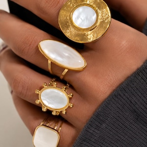 Puede incluir: Un conjunto de cuatro anillos dorados con incrustaciones de nácar blanco. El anillo superior es grande y circular con un acabado texturizado, seguido de un anillo ovalado alargado, un anillo ovalado más pequeño con cuentas y un anillo cuadrado.