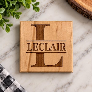 May include: A square light brown wooden coaster featuring a dark brown laser-engraved design. The design consists of a large capital letter L in the background with the name LECLAIR centered horizontally across the letter in a serif font.