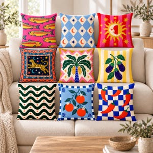 May include: A collection of nine square throw pillows featuring diverse patterns including fish on pink, blue diamonds with moons, a sun face, a leopard, a palm tree, eggplants, wavy lines with dots, oranges, and red chili peppers on a blue checkerboard.