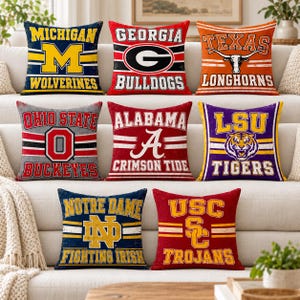 May include: A collection of eight square throw pillows featuring various college sports team logos and names, including Michigan Wolverines, Georgia Bulldogs, Texas Longhorns, Ohio State Buckeyes, Alabama Crimson Tide, LSU Tigers, Notre Dame Fighting Irish, and USC Trojans.